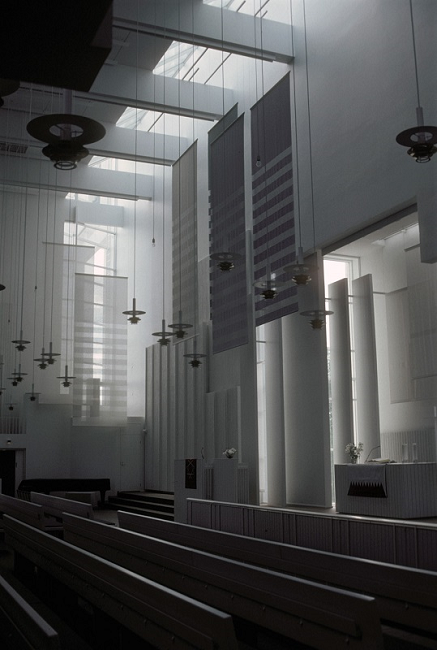It is high up to the ceiling, almost all white inside the church. Several lamps are hanging in long wires from the ceiling. At the bottom, there are rows of benches. The sunlight also seems to be shining through several places in the church.