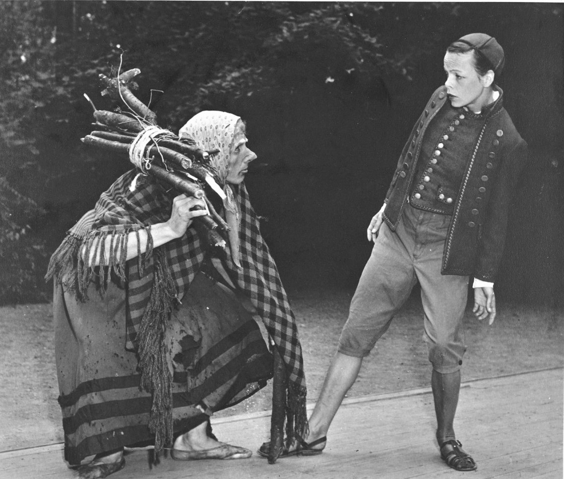 Black and white photograph of two dancers in roleplay. One male dancer dressed as a very bent old woman in old fashioned poor clothes with a bundle of wood on her shoulder. One female dancers dressed as a boy with old-fashioned costume including trousers 