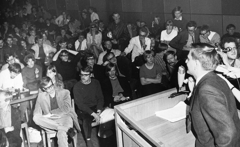 Male speakers at podium facing away into a crowd of young people sitting on chairs.