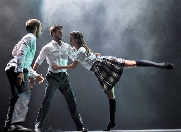 Two male(in white shirts and dark trousers) and one female dancer (in a kilt) with her leg out behind her.on a stage.