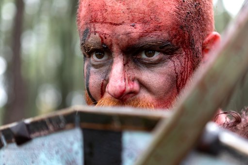A picture of a man with a red-painted face behind a shield and sword. He is only visible from above the mouth. In addition to the red blood-like paint, he has black stripes on the right half of his face.