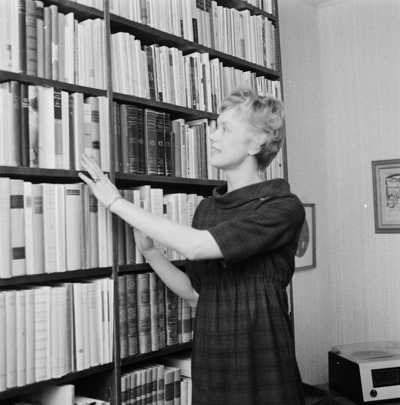 Black and white photograph of a lighthaired young woman reaching for a book from a bookshelf