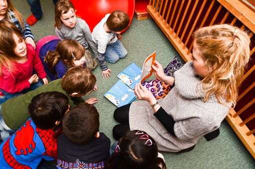 A picture of a nursery nurse sitting on the floor surrounded by kids. She is holding up a book and the kids are all looking at the front cover.