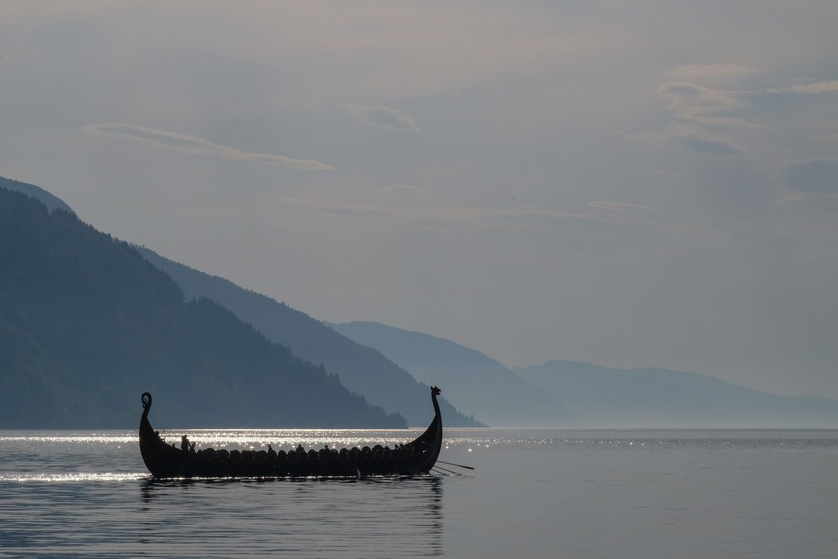 Pictured is a Viking longboat on water. The boat is mostly a silhouette against a blue background with lush mountains in different shades of blue. The sun is reflecting on the water.