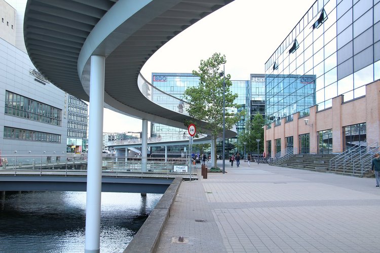 Photo taken from below Cykelslangen. A bridge that curves. Below the birdge, there is a river with small other bridges across it. Beside the Cykelslangen, there is a glass bulding.