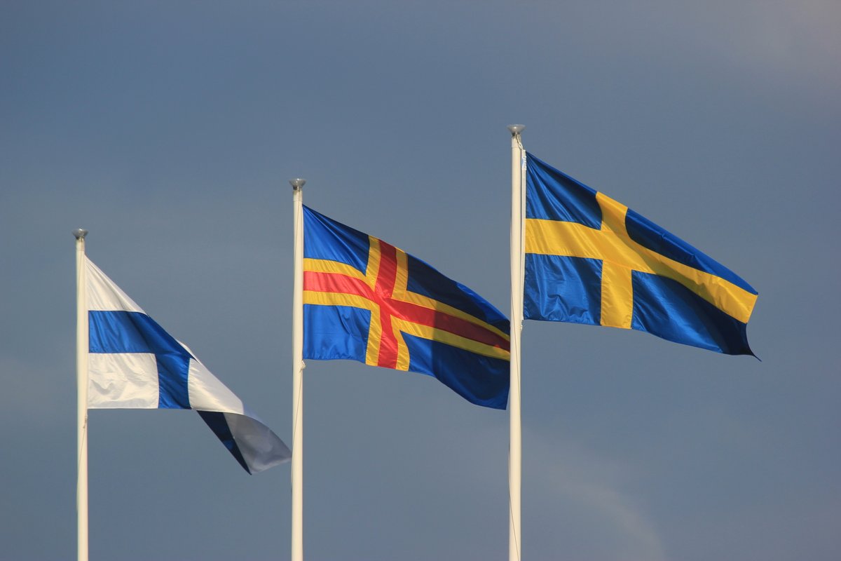 A photo of three flags besides each other. To the far left the Finnish, then the Åland flag, and to the right to Swedish.