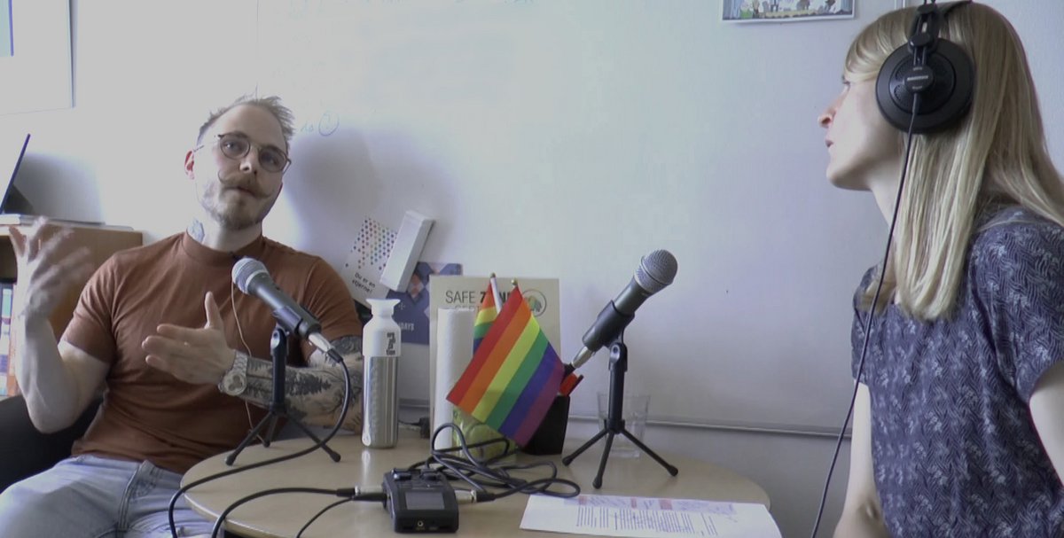 Two people talking across a table with microphones and a Pride flag.