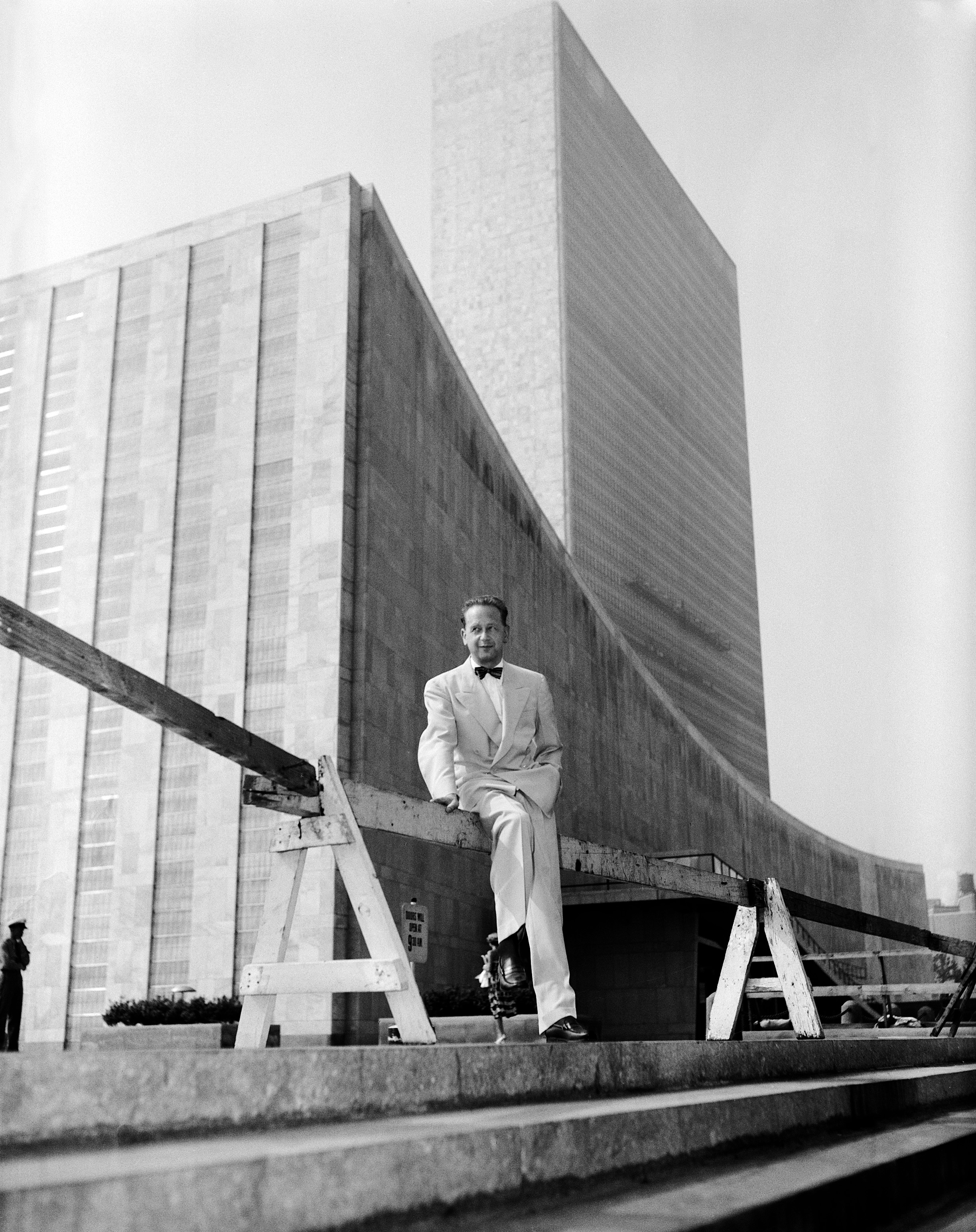 A man posing and sitting in  front of a large building with glass windows (UN headquarters)