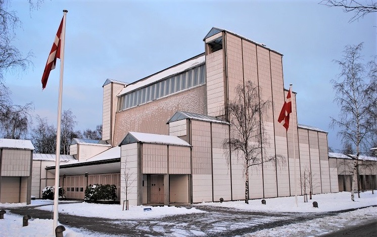 A large white building that is tall at on point but then tapers down to a more wide form. In front, there are two Danish flags. It is winter.