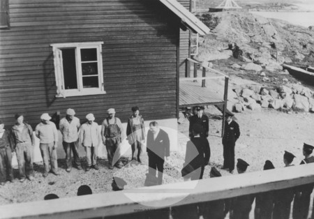 People standing in line outside a house. People on the right are wearing miliary uniforms