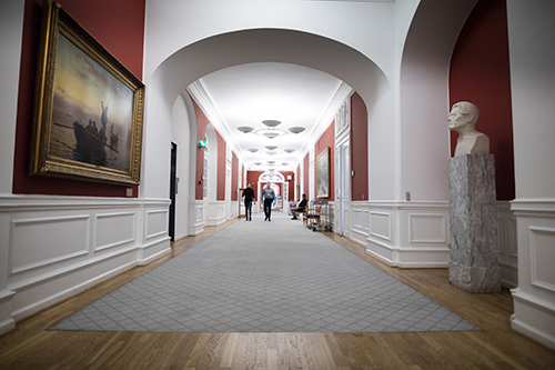 Grand-looking corridor with busts and pictures on the walls with two men walking down it.
