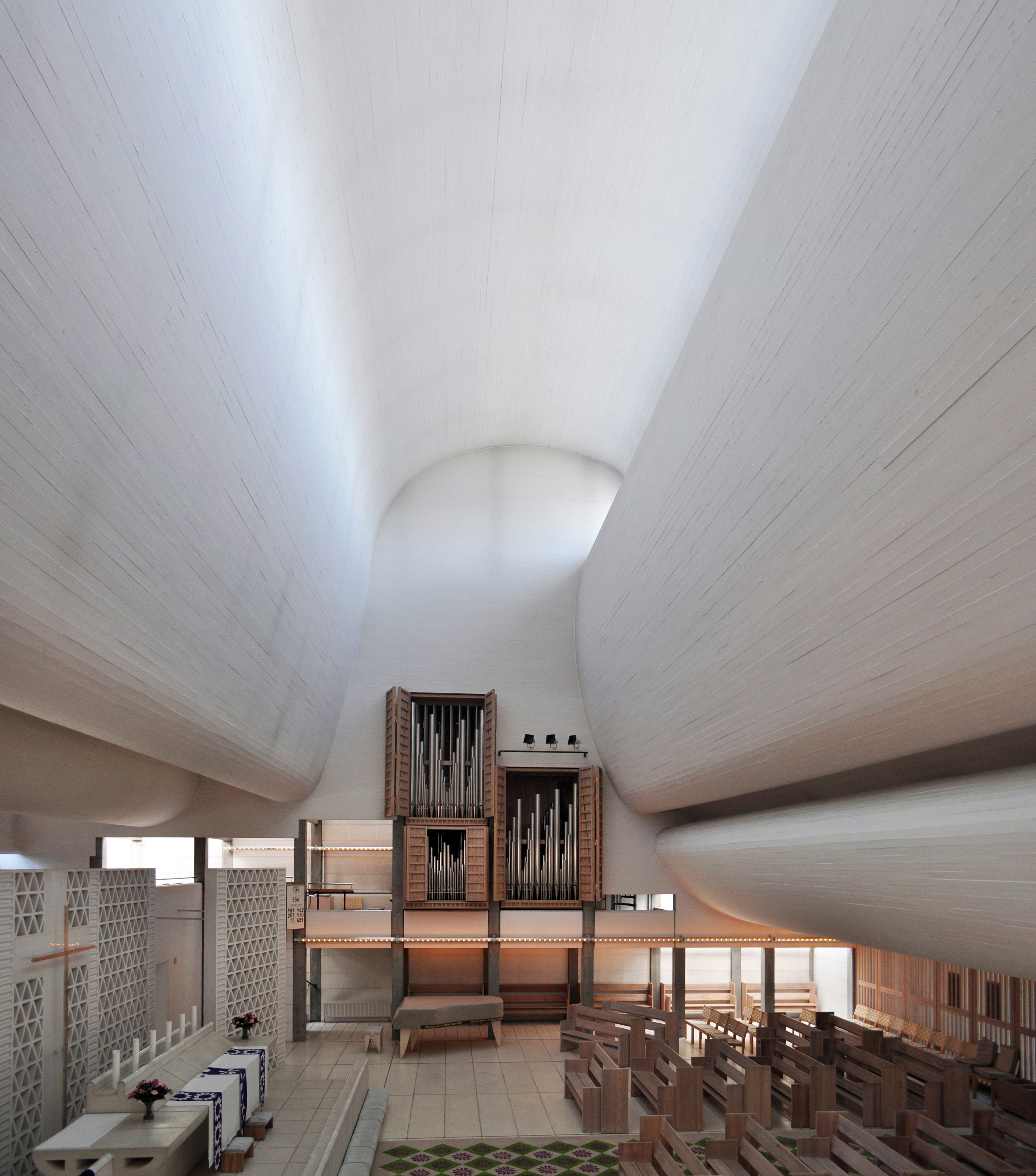 Inside of a church/a chapel, very white coloured. Looks quite closed with not much space since the walls on the sides are arching in.At the back theres an organ. There are also rows of benches to sit.