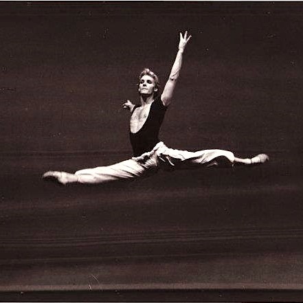 black and white picture of man leaping into the air with his left leg straight out behind him and his right straight out in front with upper body very straight and arms in a graceful ballet position