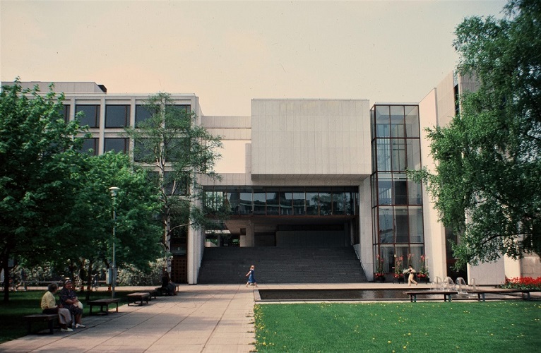 A light grey wide building with rows of square windows on its left side. In front of it is a sidewalk and some grass. There are benches on the side where two people are sitting.
