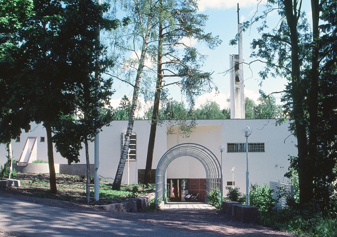 Taken outside a white church that is square shaped. It has an elegant entrance where visitors can walk through. Outside the church, there are different kinds of trees.