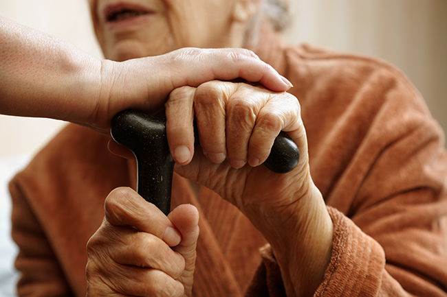 An elderly holding a cane with another person's hand is placed on top of the elderly's