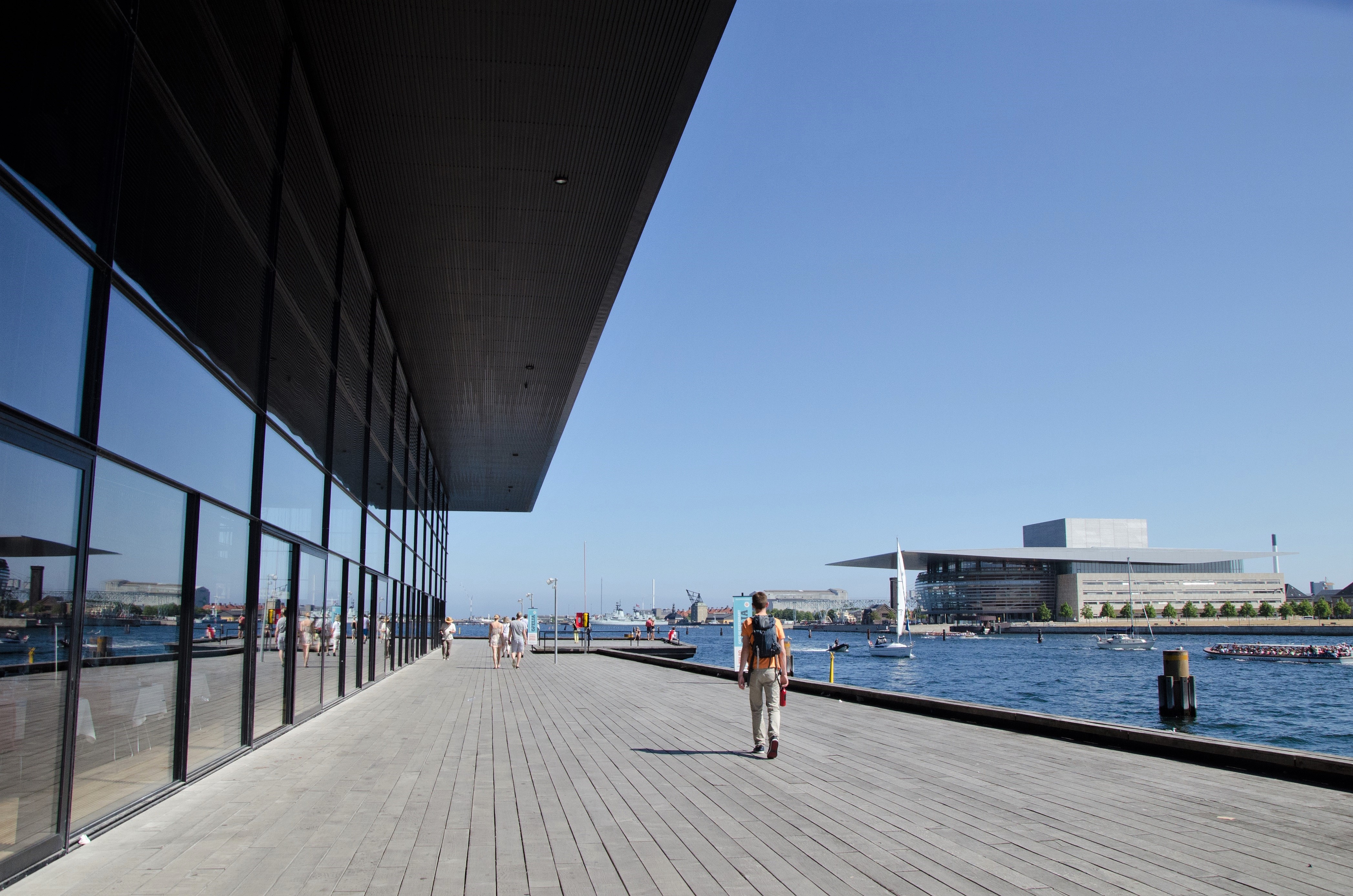 A view of the Opera House from a distance. In front of the opera house, there is the harbour. There are people walking around. It looks like summer.