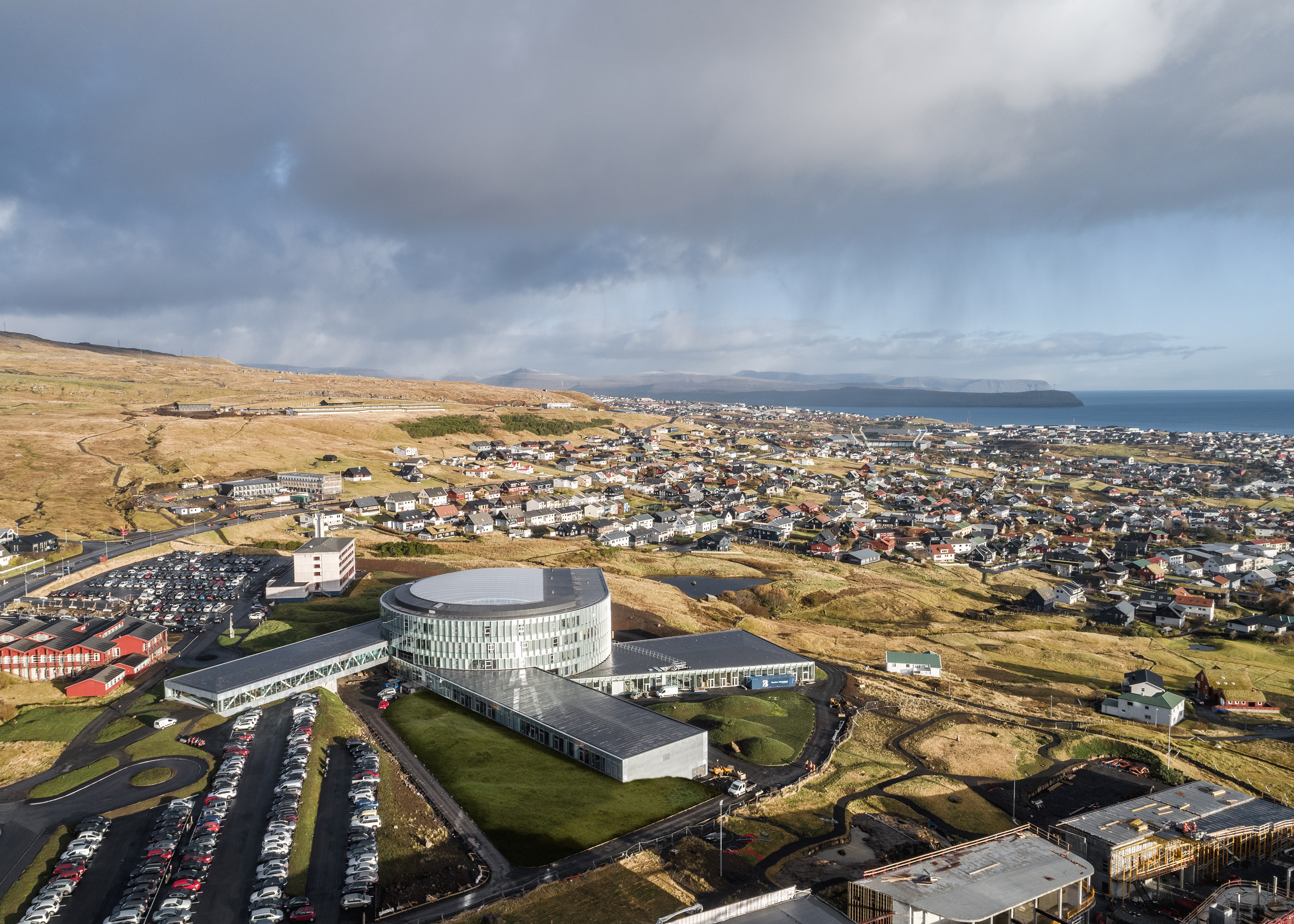 Picture taken from up high, propably a helicopter. Shows the city of Tórshavn and Glasir. The Glasir building is round shaped with glass windows all around. Three rectangular buildings are then connected all around the building.