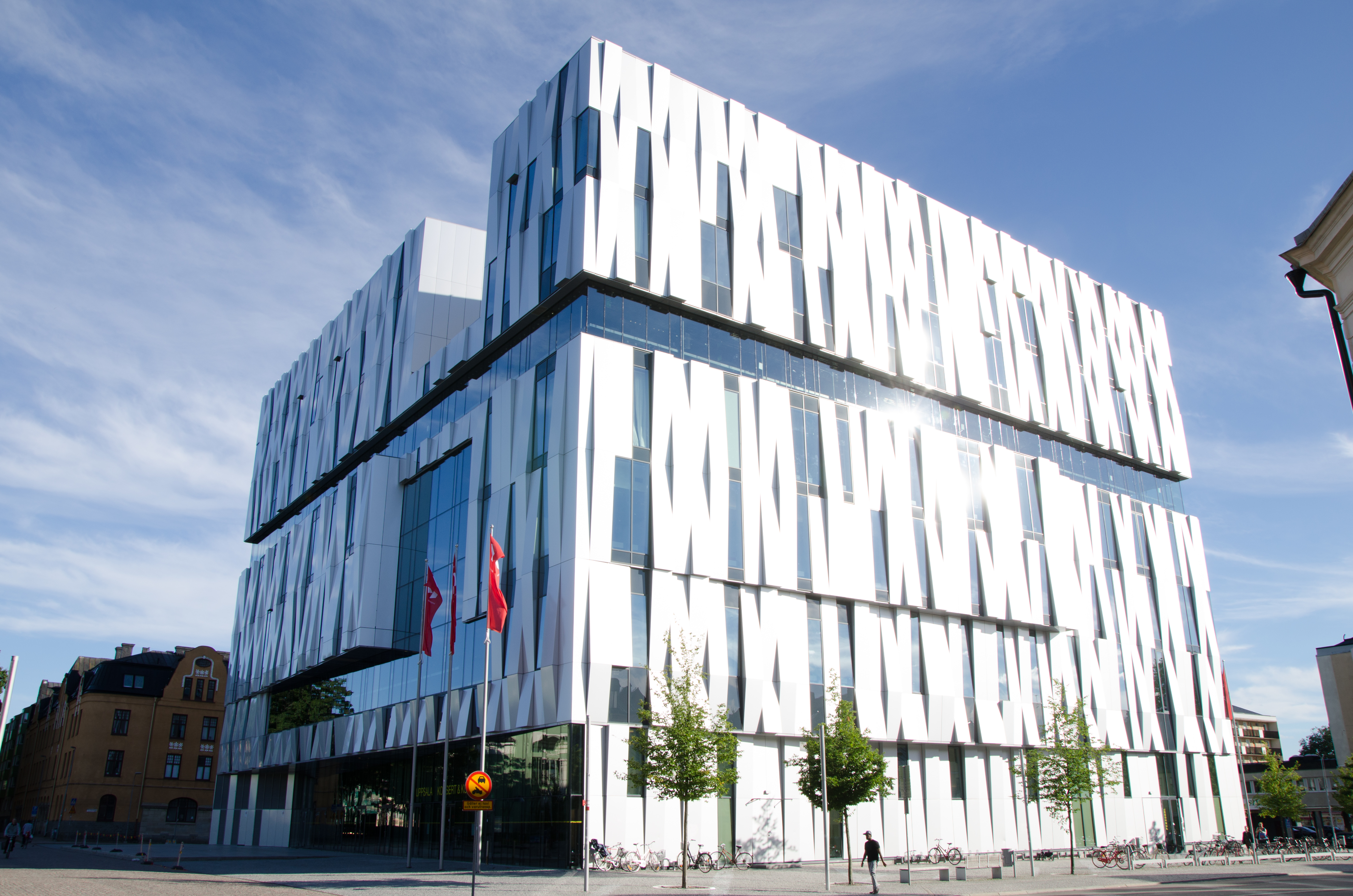 Tall white building in a criss-cross like pattern. It consists of white plates and clear windows. Around the building, there are trees planted.