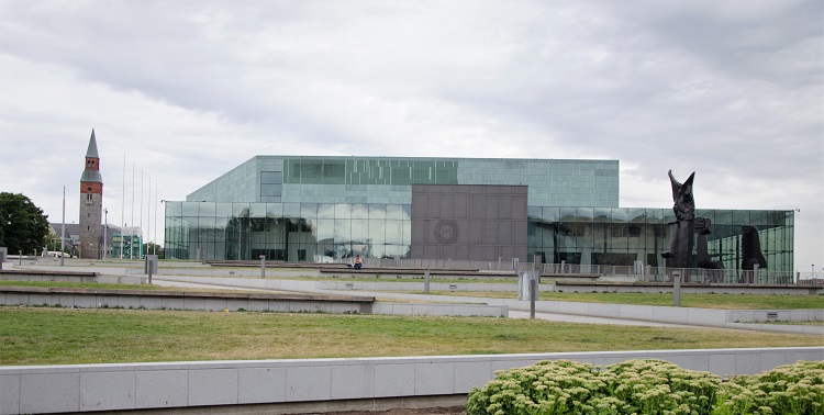 A long rektangular horisontal building in greenish glass. Beside the building, there is a long tall tower.