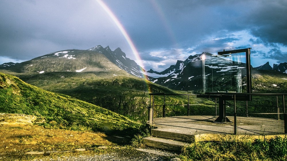 Placed up in the mountains, surrounded by green grass. Far away, you can see a tall mountain with a little snow on it. In the corner, there is a spot where people can stand and use the glass telescope (a device made of square glass) to look at the view. 