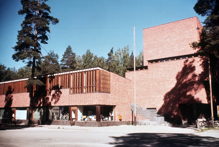 Older photo from 1952, a building of red bricks formed half wide (on the left side) and tall (on the right side). It is sunny.