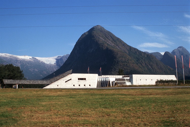 Up in the mountains on a grass field. Picture taken from afar. You can see a very long, not tall bulding that is formed like a plane without the wings. It is white. 