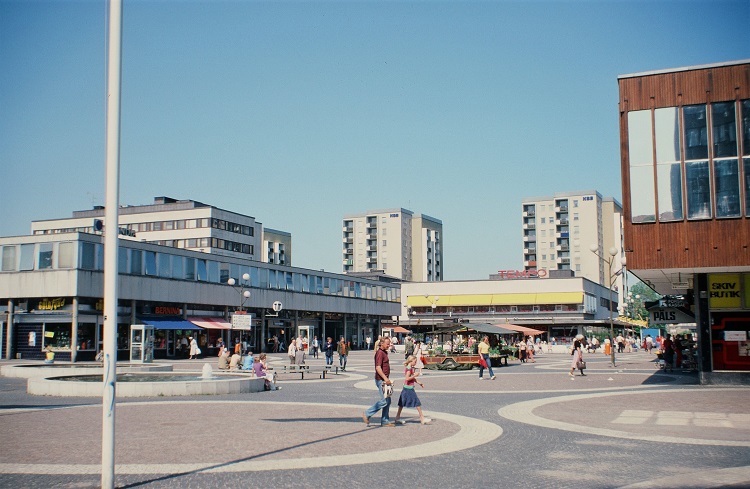 A city square with lots of people walking around enjoying themselves. In the background, there are different buildings, most of them are white except the one on the right that is red-brown with long windows