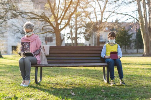 A bench in a park with green gass. The sun is shining. An old lady is sitting on the very left side of the bench wearing a face mask, and on the very right is a small boy also wearing a facemask. She is reading a book, he is looking at a tablet.