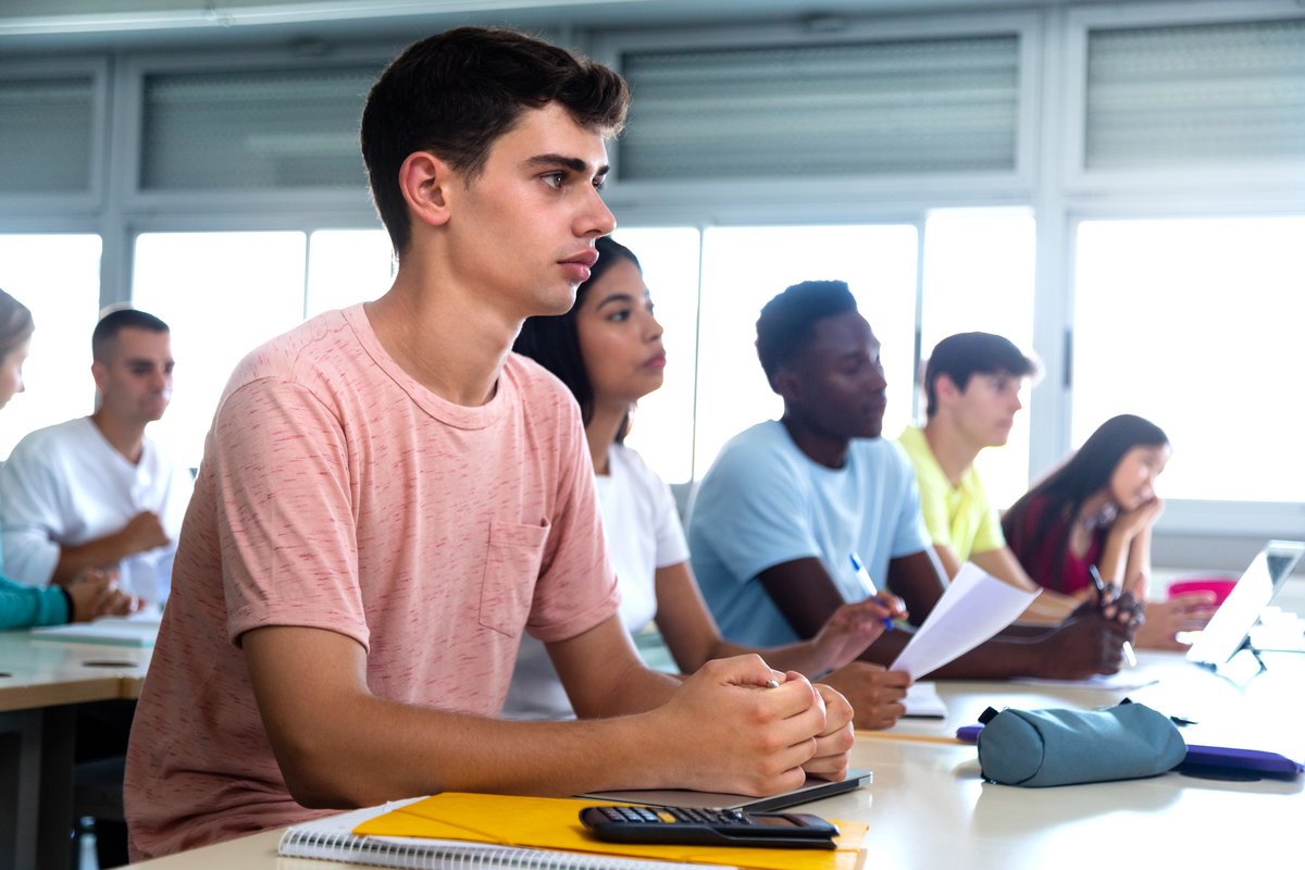 A picture of a white boy with dark brown hair and a pink shirt sitting in a classroom. The picture is taken from a side angle and sitting beside the boy are a brown girl and a black boy. In the blurred background are another white boy and a white girl.