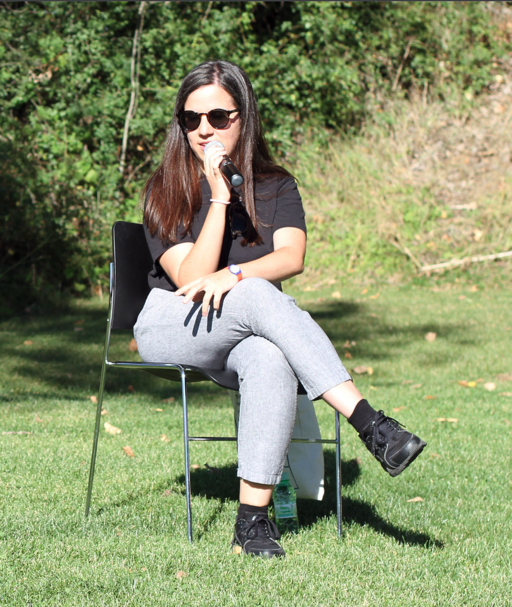 A photo of a woman sitting on a chair outside on the grass. Her legs are crossed. One hand is resting on her thigh, the other is holding a microphone. The sun is shining on her and she is wearing sunglasses. She is wearing jeans and a black t-shirt. Her hair is long and brown.