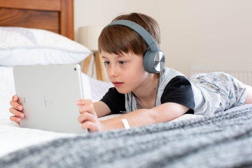 Pictured is a young boy is laying on a bed, wearing headphones and looking at an iPad he is holding in his hands. The photo is very bright, and the boy looks very focused on whatever he is watching.
