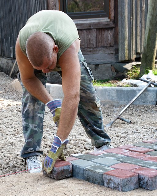 Pictured is a contemporary male, laying bricks. He is wearing cargo pants and a grey t-shirt, and looks exhausted while he is engaging in difficult, manual labour.