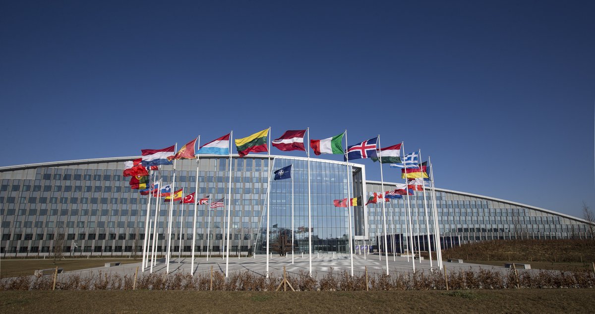 The front of the new NATO headquarters in Brussels, a modern looking building with a glass facade. In the front is a circle of flagpoles, with flags for all the membercountries.