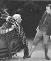 Black and white photograph of two dancers in roleplay. One male dancer dressed as a very bent old woman in old fashioned poor clothes with a bundle of wood on her shoulder. One female dancers dressed as a boy with old-fashioned costume including trousers 
