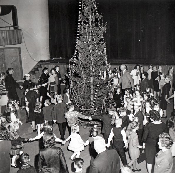 black and white photo of a christmas tree party. a crowd of people are holding hands and dancing around the christmas tree which is placed in the middle of the large circle.