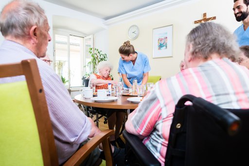 En ældre mand og en ældre kvinder sidder ved et bord på et plejehjem. På den modsatte side af bordet snakker en ældre kvinde med en ung kvindelig medarbejder.