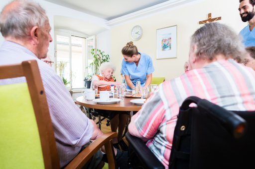 An elderly man and an elderly woman are sitting at a table in a nursing home. On the opposite side of the table, an elderly woman is talking to a young female staff member.