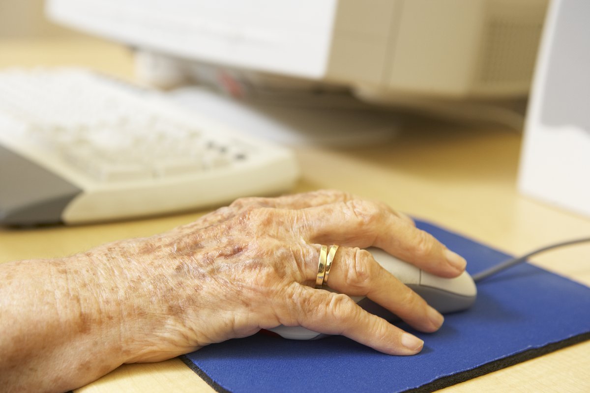 A picture of a hand holding a computer mouse on a blue mousepad. In the background is an old computer. The hand is veiny and has a wedding ring, clearly belonging to an elderly person.