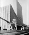 A man posing and sitting in  front of a large building with glass windows (UN headquarters)