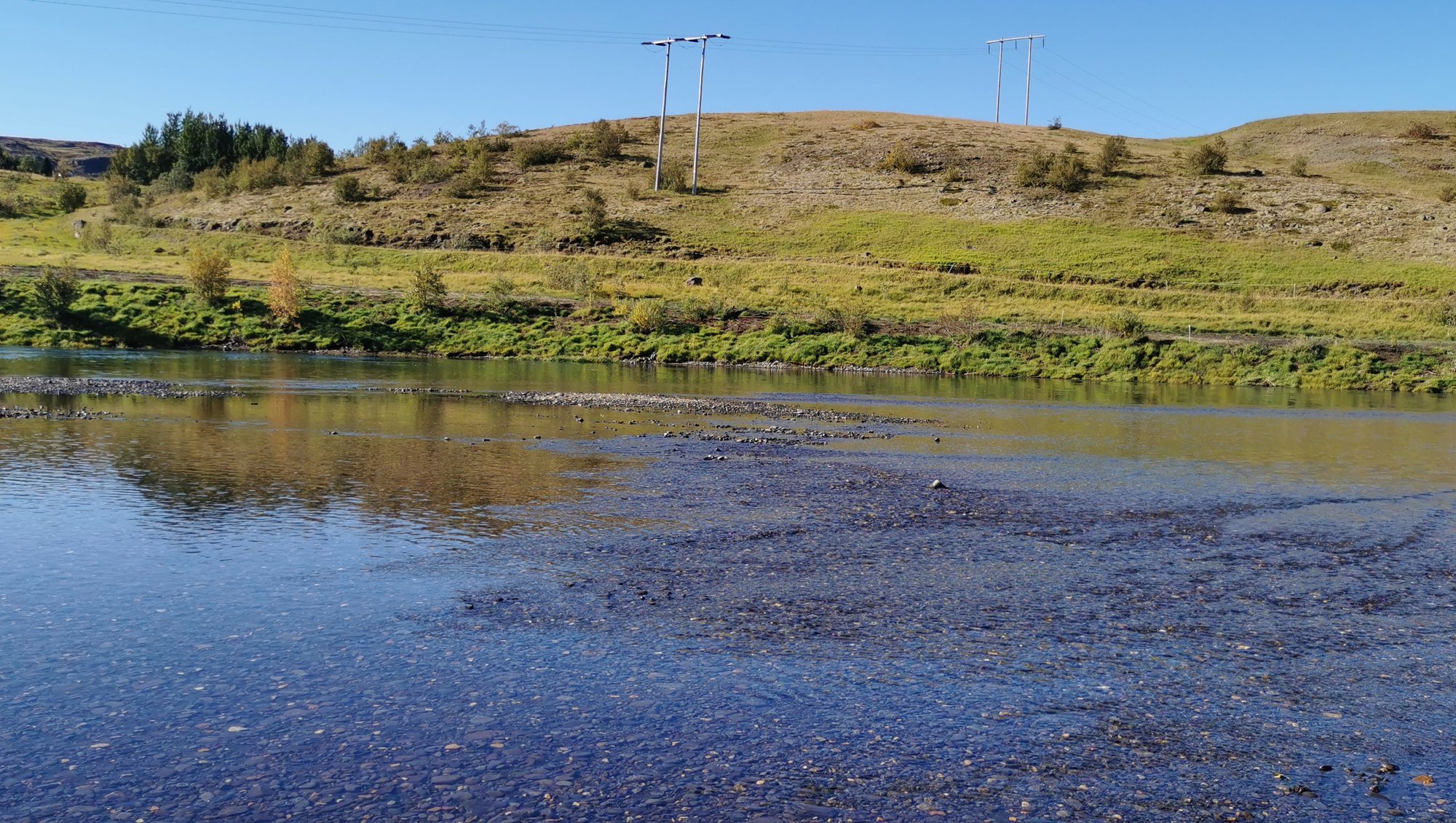 In the foreground is blue water, the restored bank. Behind it are green hills, on which a couple of power cables are hanging in the air. In the very background is the blue sky with no clouds.