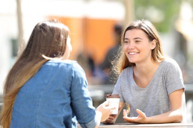 Pictured is two women sitting at a cafe table. One is facing the camera, the other has her back towards the camera. They are drinking coffee to go and smiling. The woman facing the camera is gesticulating something with once hand.