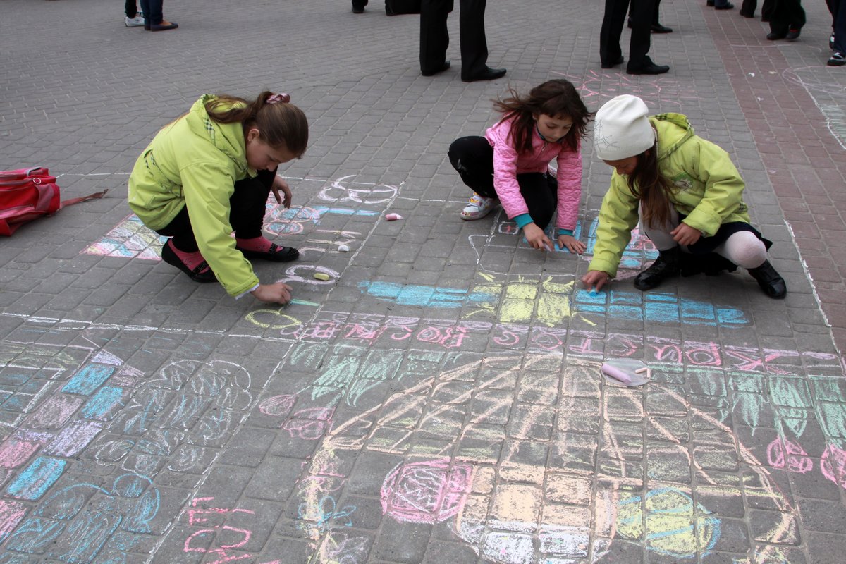 A picture of three children in winter coats, crouched on outside tiles. They are drawing with chalk in many colours on the ground. 