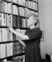 Black and white photograph of a lighthaired young woman reaching for a book from a bookshelf
