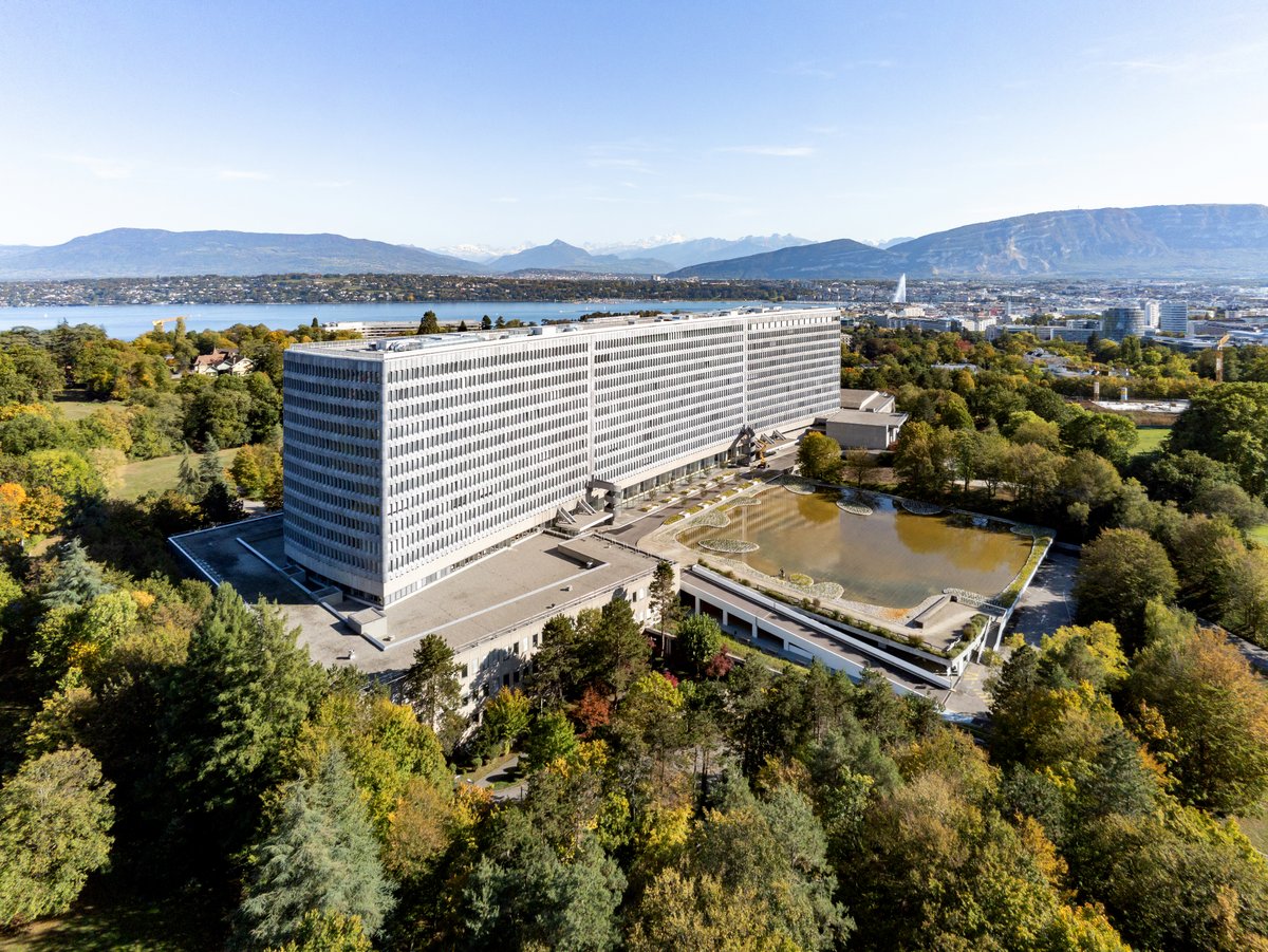 A photo from above of a white, modern building with lots of window. It is surrounded by trees. In the background are mountains.