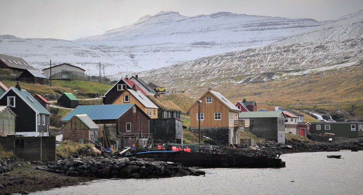 A picture of wooden houses along the waterside in different, muted colours. In the background are tall, snowty mountains.