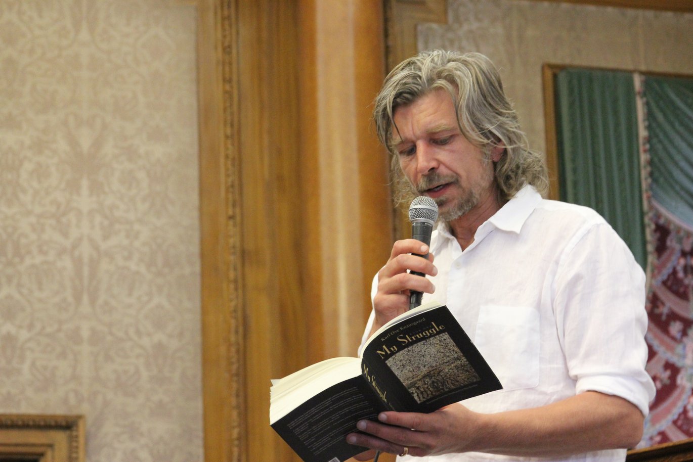 A waist-up picture of Karl Ove Knausgård. He is wearing a white shirt and has grey hair and a grey beard. In one hand he is holding a book with the title "My Struggle" and in the other hand he has a microphone lifted to his mouth.