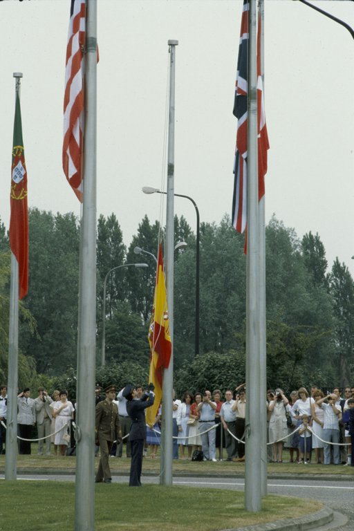 A person raising the Spanish flag during a cerenomy. Many people are observing from behind a rope-fence, some taking pictures.