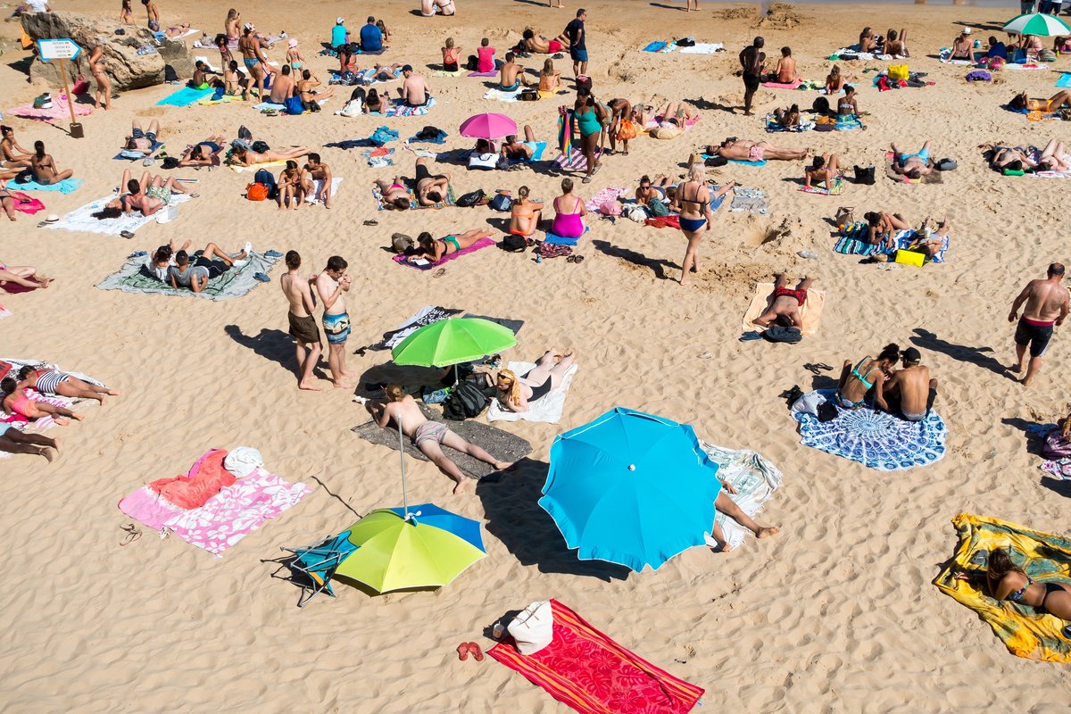 En sandstrand i strålande solsken. Det är trångt och folk solar på handdukar som har många olika färger med parasoll som också har många olika färger.