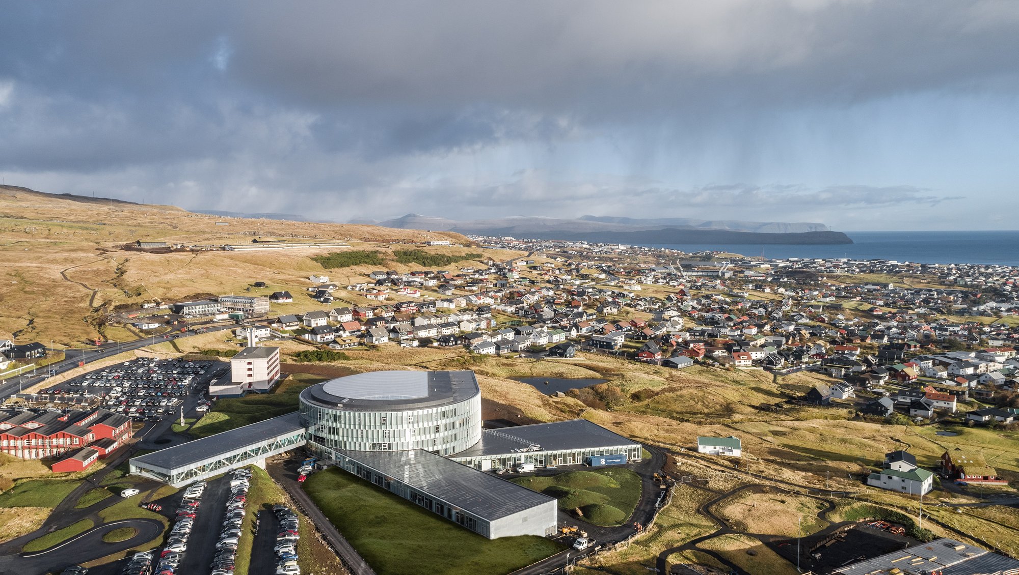 Picture taken from up high, propably a helicopter. Shows the city of Tórshavn and Glasir. The Glasir building is round shaped with glass windows all around. Three rectangular buildings are then connected all around the building.